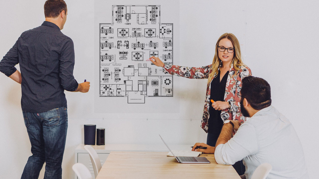 An image of one female pointing at an office design plan on the wall with two male colleagues watching